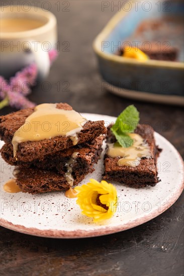 Chocolate brownie with caramel sauce with a cup of coffee on black concrete background. side view, close up, selective focus