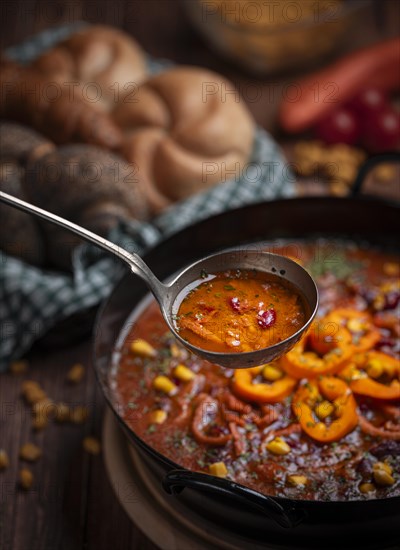 Soup ladle scooping bean soup with sweetcorn and peppers from a pot, basket with pastries in the background