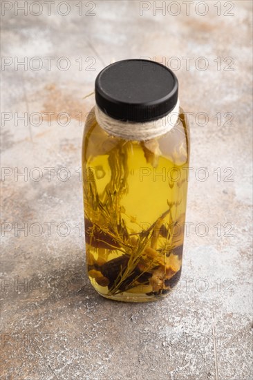 Sunflower oil in a glass jar with various herbs and spices, lavender, sesame, rosemary on a brown concrete background. Side view, close up