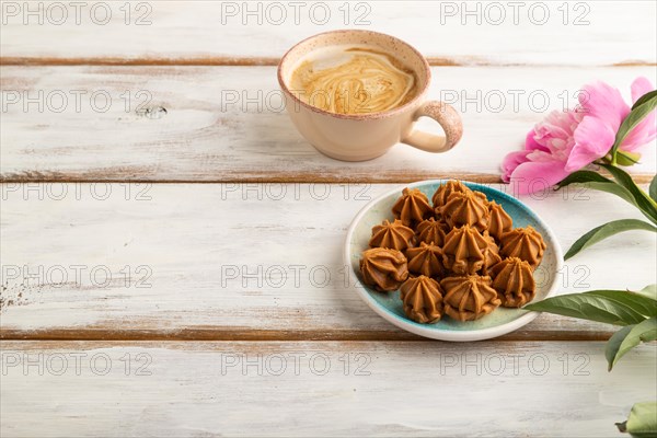 Homemade soft caramel fudge candies on blue plate and cup of coffee on white wooden background, peony flower decoration. side view, copy space