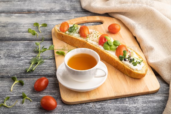Long white bread sandwich with cream cheese, tomatoes and microgreen on gray wooden background and linen textile. side view, close up