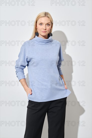 Studio portrait of classy blonde woman in blue sweater and black trousers with her hands in pockets