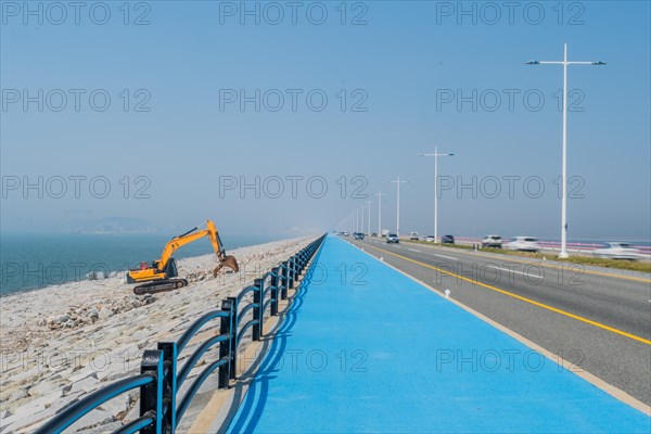 Backhoe sitting on ocean causeway next to blue concrete sidewalk on divided highway with speeding cars passing by. Motion blur caused by slow shutter speed