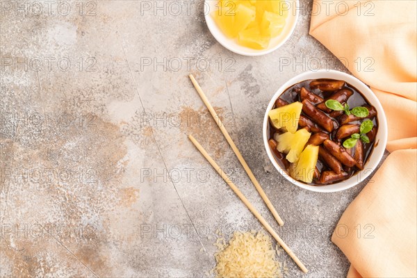 Tteokbokki or Topokki, fried rice cake stick, popular Korean street food with spicy jjajang sauce and pineapple on gray concrete background and orange textile. Top view, flat lay, copy space