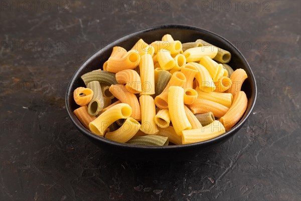 Rigatoni colored raw pasta with tomato, eggs, spices, herbs on black concrete background. Side view, close up