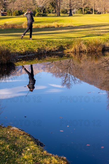 Male Golfer Reflected in a Water Pond and Hitting the Golf Ball on Fairway on Golf Course with Mountain in a Sunny Day in Switzerland
