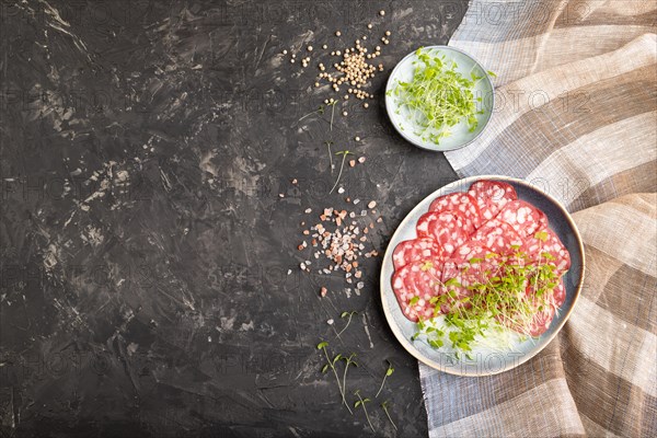 Slices of smoked cervelat salami sausage with spinach microgreen, salt and pepper on black concrete background and beige textile. Top view, flat lay, copy space