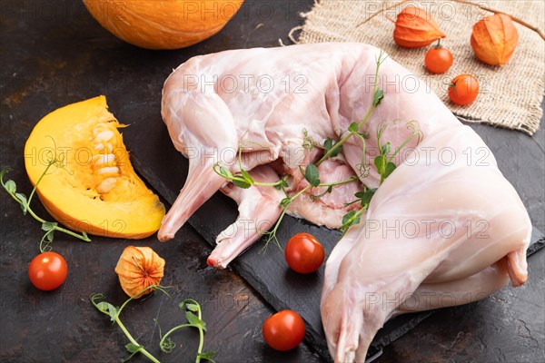 Whole raw rabbit with pumpkin, tomatoes, pea sprouts on a black concrete background and linen textile. Side view, close up