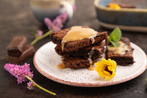 Chocolate brownie with caramel sauce with a cup of coffee on black concrete background. side view, close up, selective focus