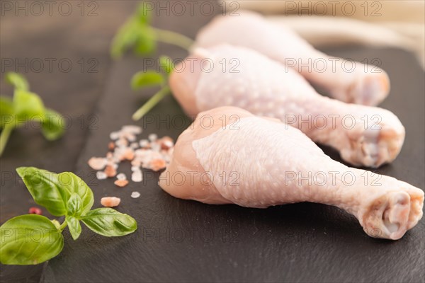 Raw chicken legs with herbs and spices on a black slate cutting board on a black concrete background and linen textile. Side view, close up, selective focus