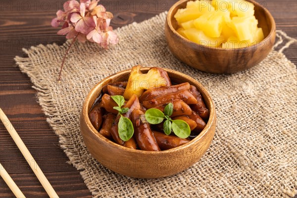 Tteokbokki or Topokki, fried rice cake stick, popular Korean street food with spicy jjajang sauce and pineapple on brown wooden background and linen textile. Side view