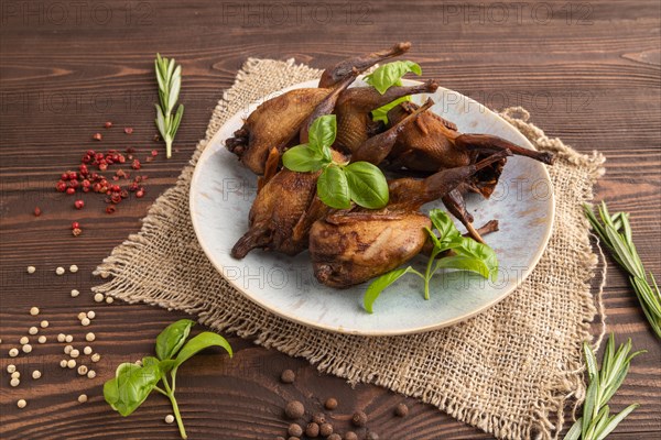Smoked quails with herbs and spices on a ceramic plate with linen textile on a brown wooden background. Side view, close up, selective focus