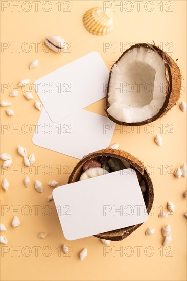 White paper business card with coconut and seashells on orange pastel background. Top view, flat lay, copy space. Tropical, healthy food, vacation, holidays concept