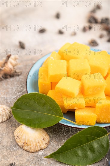 Dried and candied mango cubes on blue plate on brown concrete background and linen textile. Side view, close up, selective focus, vegan, vegetarian food concept