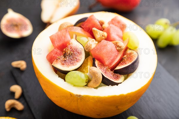 Vegetarian fruit salad of watermelon, grapes, figs, pear, orange, cashew on slate board on a black concrete background. Side view, close up, selective focus