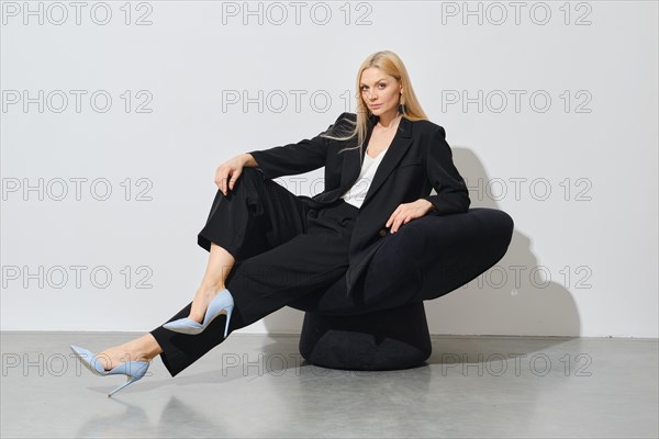 Modern successful businesswoman in black pantsuit sits on black bean bag indoors