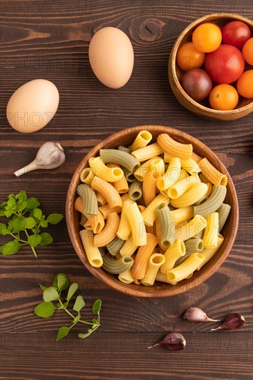 Rigatoni colored raw pasta with tomato, eggs, spices, herbs on brown wooden background. Top view, flat lay, close up