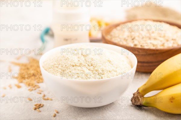 Powdered milk and oatmeal, banana baby food mix, infant formula, pacifier, bottle, spoon on gray concrete background. Side view, close up, selective focus, artificial feeding concept