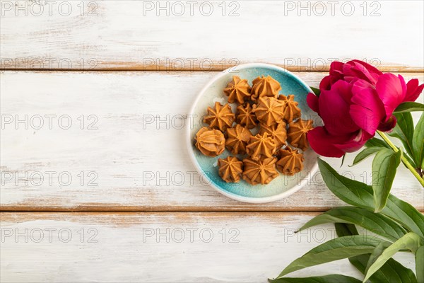 Homemade soft caramel fudge candies on blue plate on white wooden background, peony flower decoration. top view, flat lay, copy space