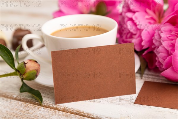 Brown business card with pink peony flowers and cup of coffee on white wooden background. side view, copy space, still life. Breakfast, morning, spring concept