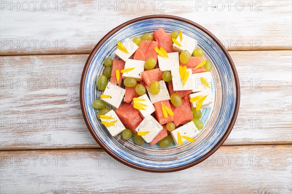 Vegetarian salad with watermelon, feta cheese, and grapes on blue ceramic plate on white wooden background. top view, close up, flat lay