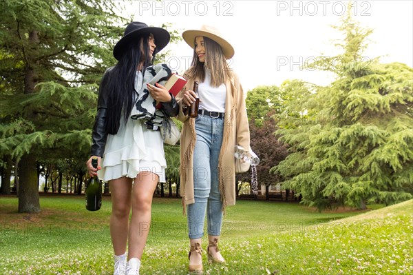 Front view of a lesbian couple walking in the park with a bottle of champagne and glasses to celebrate their courtship