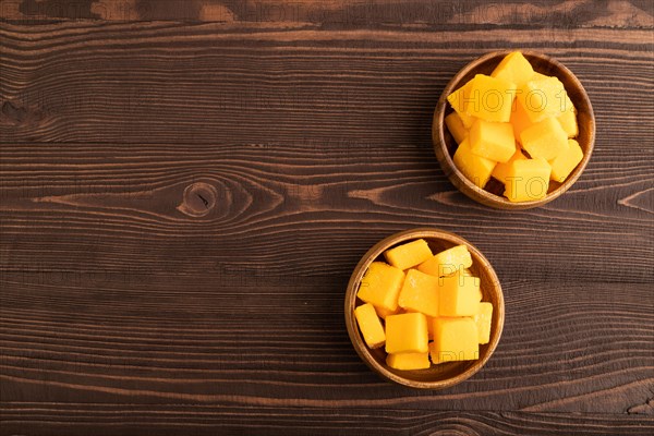 Dried and candied mango cubes in wooden bowls on brown wooden textured background. Top view, flat lay, copy space, vegan, vegetarian food concept