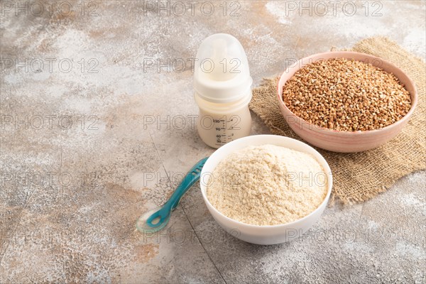 Powdered milk and buckwheat baby food mix, infant formula, pacifier, bottle, spoon on brown concrete background and linen textile. Side view, copy space, artificial feeding concept
