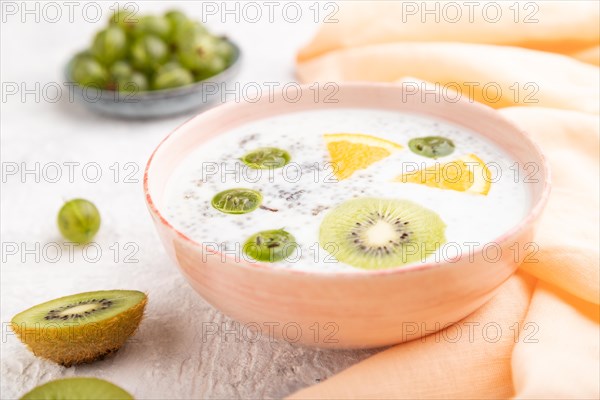 Yogurt with kiwi, gooseberry, chia in ceramic bowl on gray concrete background and orange linen textile. Side view, close up, selective focus