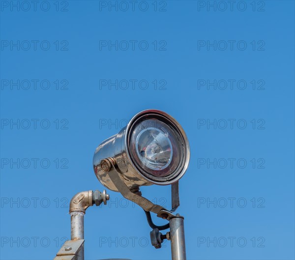 Searchlight with cracked lens mounted on pole against blue sky