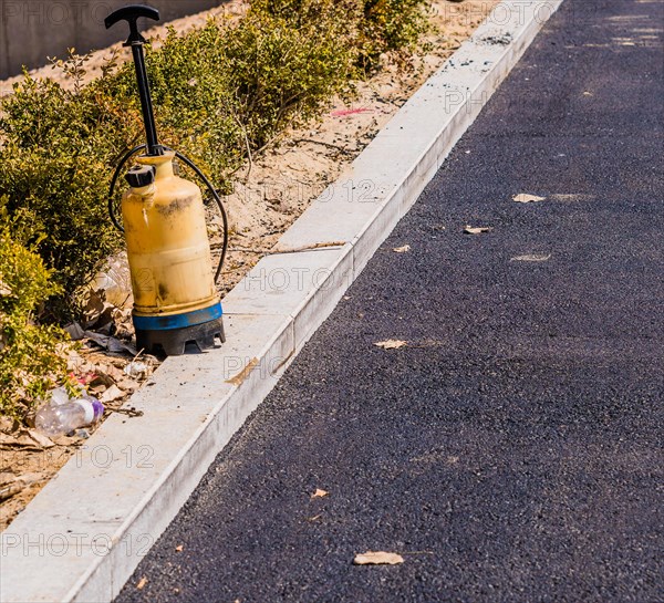 Plastic industrial spray bottle sitting on the curb of parking lot with black asphalt paving and a row of green bushes