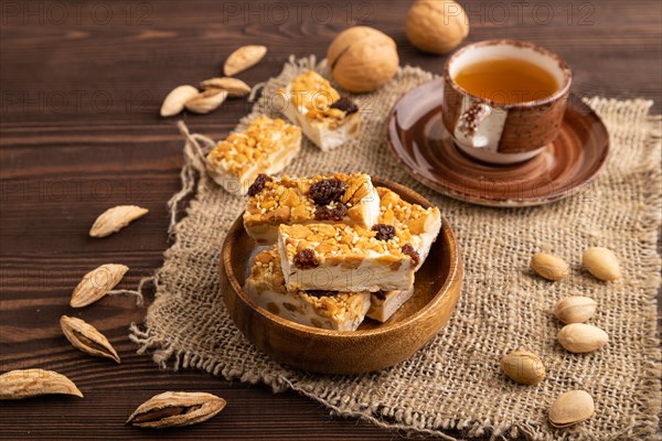 Traditional candy nougat with nuts and sesame with cup of green tea on brown wooden background and linen textile. side view, close up