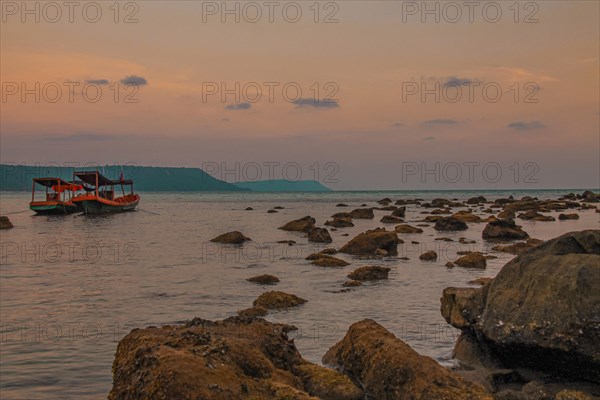 Twilight over a rocky shoreline with anchored boats and a calm sea under a soft orange sky. Koh Rong, Cambodia, Asia