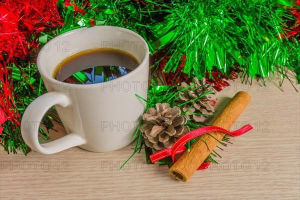 Cup of black coffee in a white cup next to pine cones and cinnamon stick with red and green Christmas tensile in background reflecting in coffee