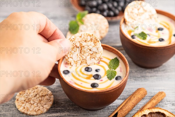 Yoghurt with bilberry and caramel in clay bowl on gray wooden, hand dipping cookies in yogurt, side view, close up, selective focus