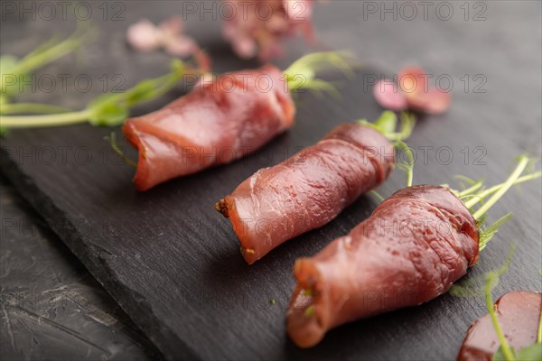 Slices of smoked salted meat with green pea microgreen on black concrete background. Side view, close up, selective focus