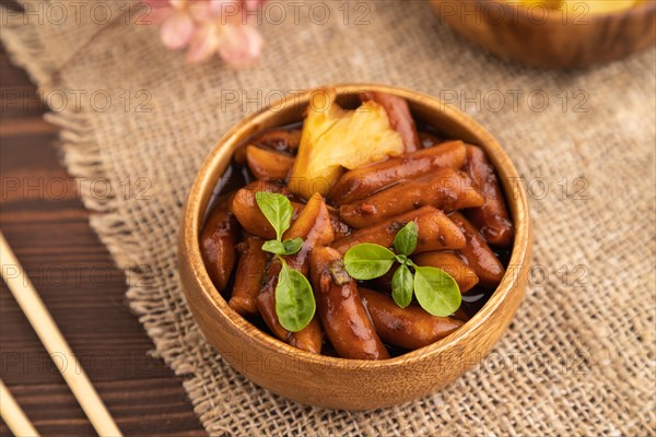 Tteokbokki or Topokki, fried rice cake stick, popular Korean street food with spicy jjajang sauce and pineapple on brown wooden background and linen textile. Side view, selective focus