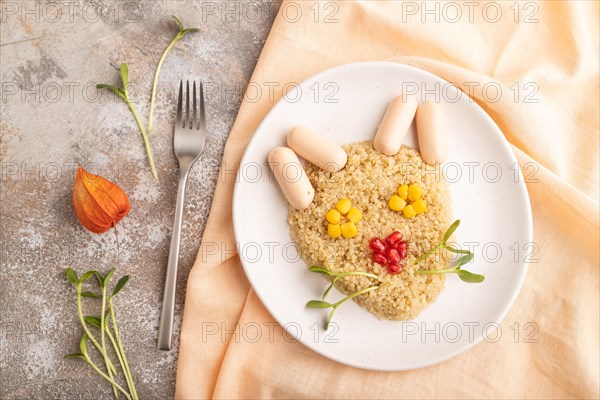 Funny mixed quinoa porridge, sweet corn, pomegranate seeds and small sausages in form of cat face on brown concrete background and orange textile. Top view, flat lay. Food for children, healthy food concept