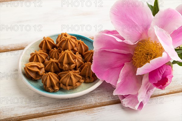 Homemade soft caramel fudge candies on blue plate on white wooden background, peony flower decoration. side view, close up