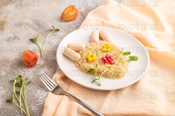 Funny mixed quinoa porridge, sweet corn, pomegranate seeds and small sausages in form of cat face on brown concrete background and orange textile. Side view, food for children, healthy food concept