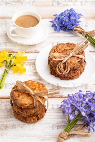 Oatmeal cookies with spring snowdrop flowers bluebells, narcissus and cup of coffee on white wooden background. side view, close up, still life. Breakfast, morning, spring concept