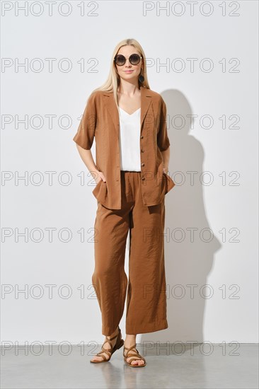 Cheerful blonde woman showcases a modern summer casual outfit. The loose brown suit and white inner top are paired with leather sandals