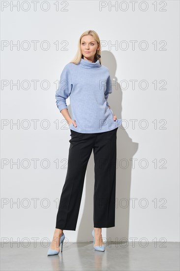A modern fashion model posing with hands in pockets of classic black trousers wearing a stylish blue sweater and stilettos
