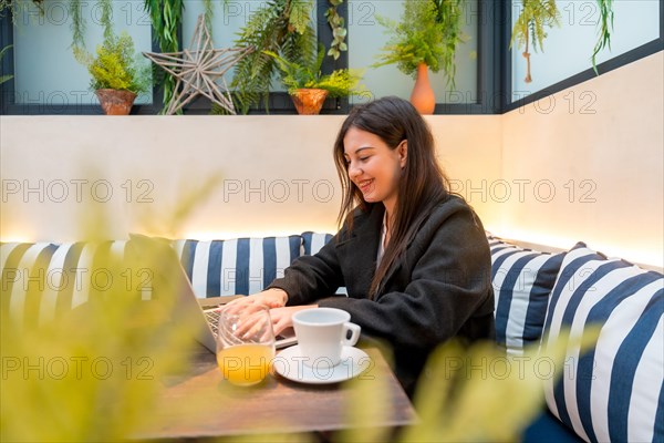 Smiling woman using laptop sitting in a colorful cafeteria drinking orange juice and coffee