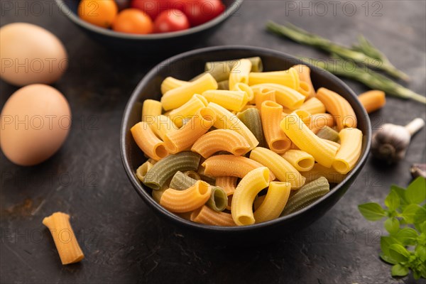 Rigatoni colored raw pasta with tomato, eggs, spices, herbs on black concrete background. Side view, close up, selective focus