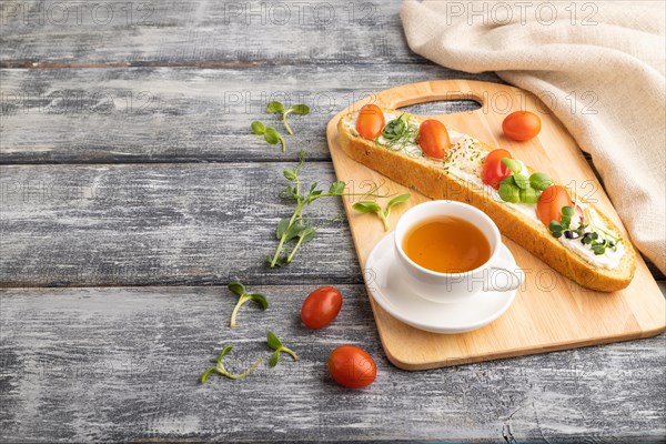 Long white bread sandwich with cream cheese, tomatoes and microgreen on gray wooden background and linen textile. side view, copy space