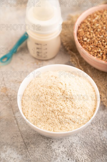Powdered milk and buckwheat baby food mix, infant formula, pacifier, bottle, spoon on brown concrete background and linen textile. Side view, close up, selective focus, artificial feeding concept