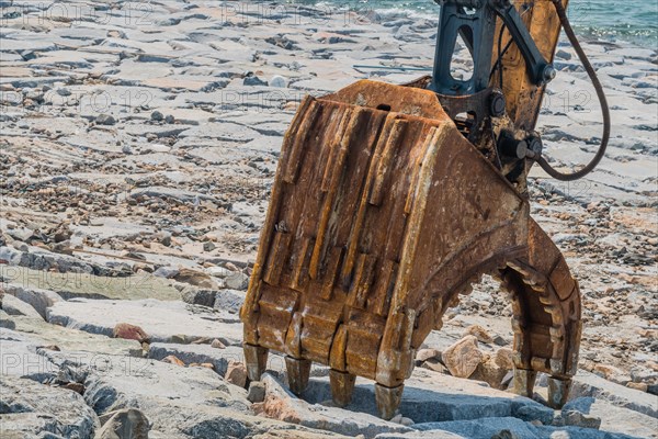 Hydraulic jaws attached to arm of backhoe in open position resting on rocky shore of man made dike