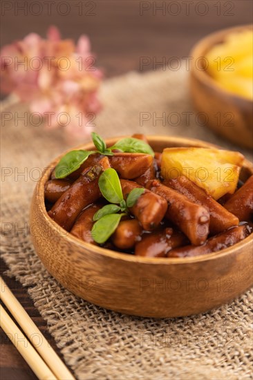 Tteokbokki or Topokki, fried rice cake stick, popular Korean street food with spicy jjajang sauce and pineapple on brown wooden background and linen textile. Side view, selective focus