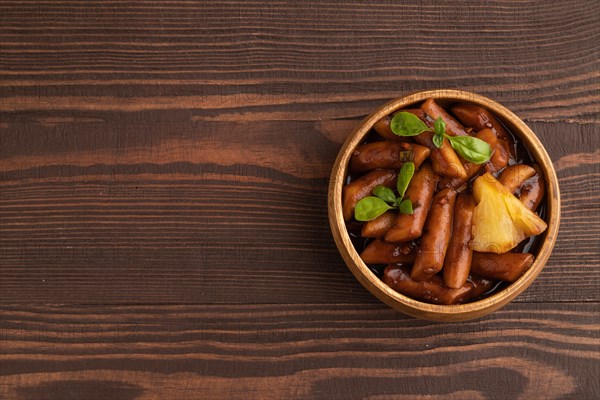 Tteokbokki or Topokki, fried rice cake stick, popular Korean street food with spicy jjajang sauce and pineapple on brown wooden background. Top view, flat lay, copy space